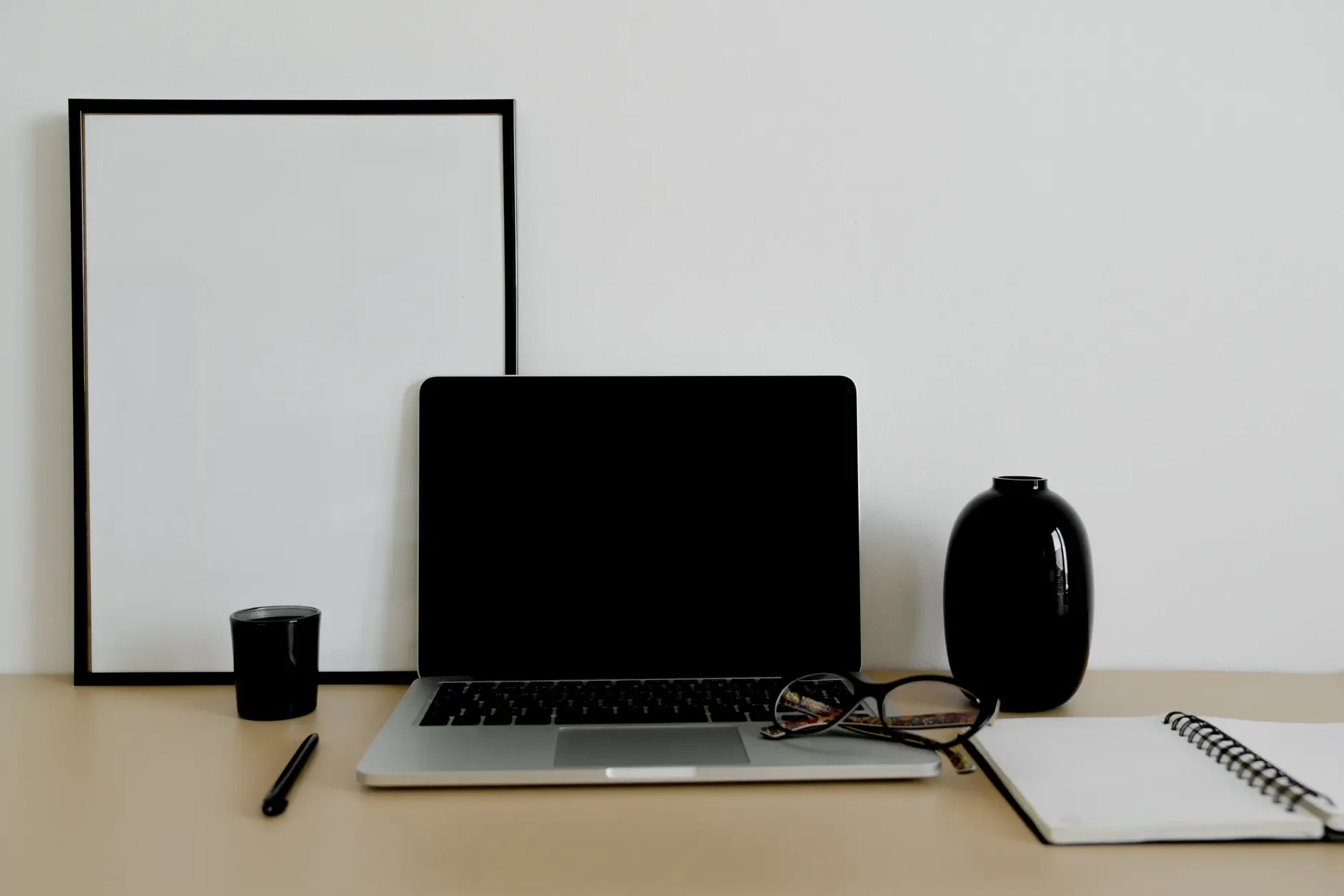 Laptop mit dunklem Bildschirm, Brille, offenem Spiralnotizbuch und Stift auf einem aufgeräumten Schreibtisch.
